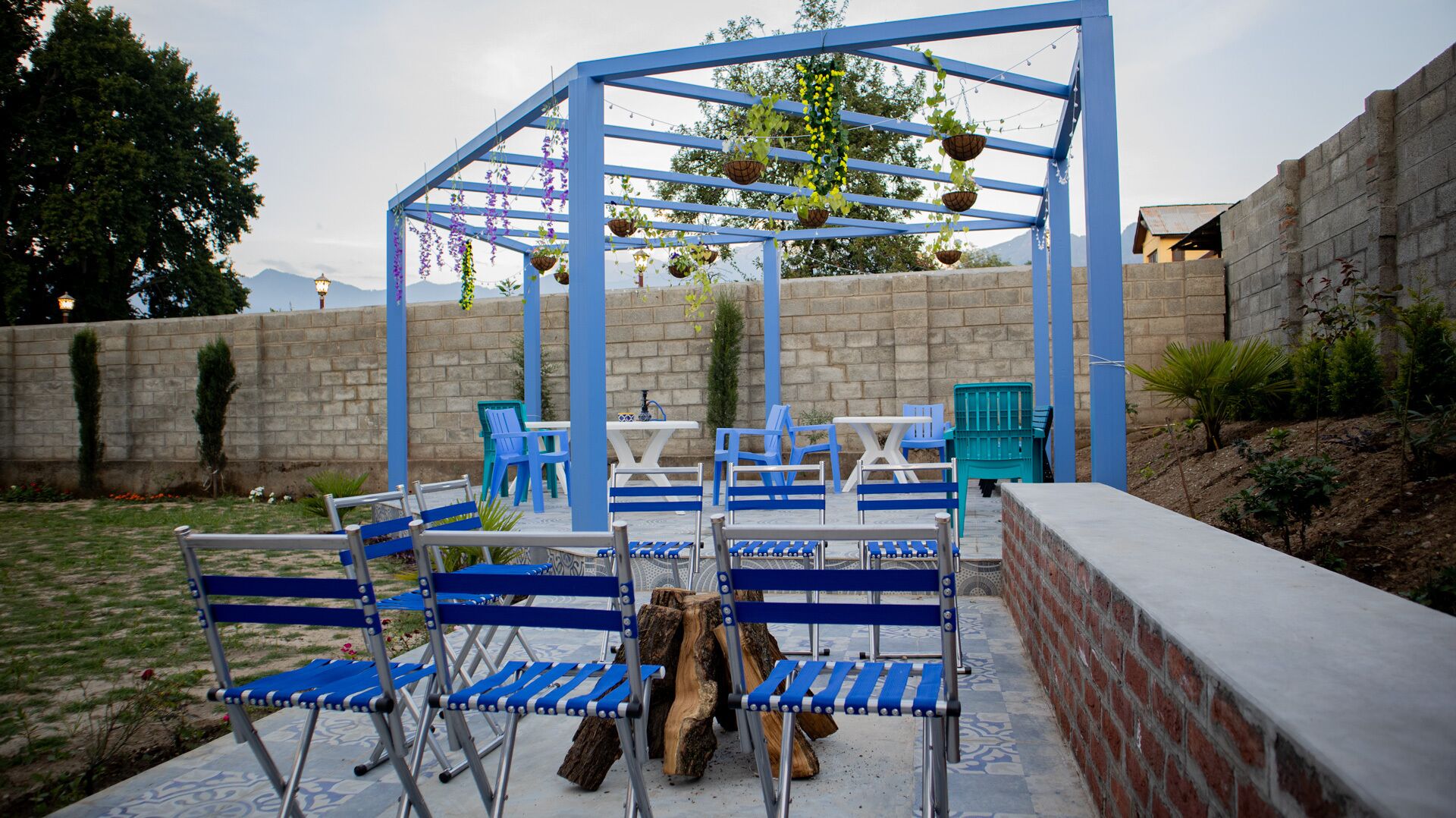 An outdoor seating area with blue chairs and tables, a pergola with hanging plants, and a brick wall in the background.
