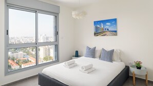 A modern hotel room with a large window, a bed with neatly folded towels, a city view, and a painting on the wall. - MAMAD Deluxe Apt & Terrace with City Overview by FeelHome (Holon)