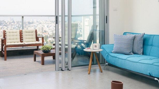 A modern living room with a blue sofa, a wooden chair, a small table with a plant, and a view of the city through large glass doors. - MAMAD Deluxe Apt & Terrace with City Overview by FeelHome (Holon)