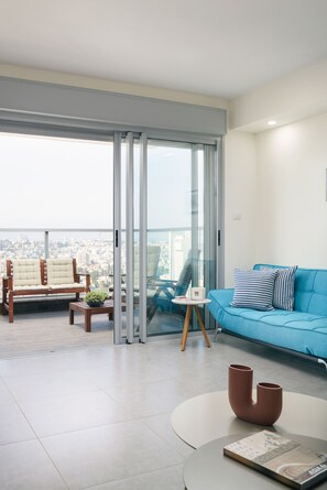 A modern living room with a blue sofa, a wooden chair, a small table with a plant, and a view of the city through large glass doors. - MAMAD Deluxe Apt & Terrace with City Overview by FeelHome (Holon)