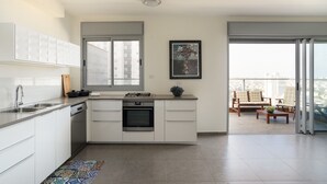 A modern kitchen with white cabinets, a gas stove, and a dishwasher. There is a large window and a balcony with outdoor furniture. - MAMAD Deluxe Apt & Terrace with City Overview by FeelHome (Holon)
