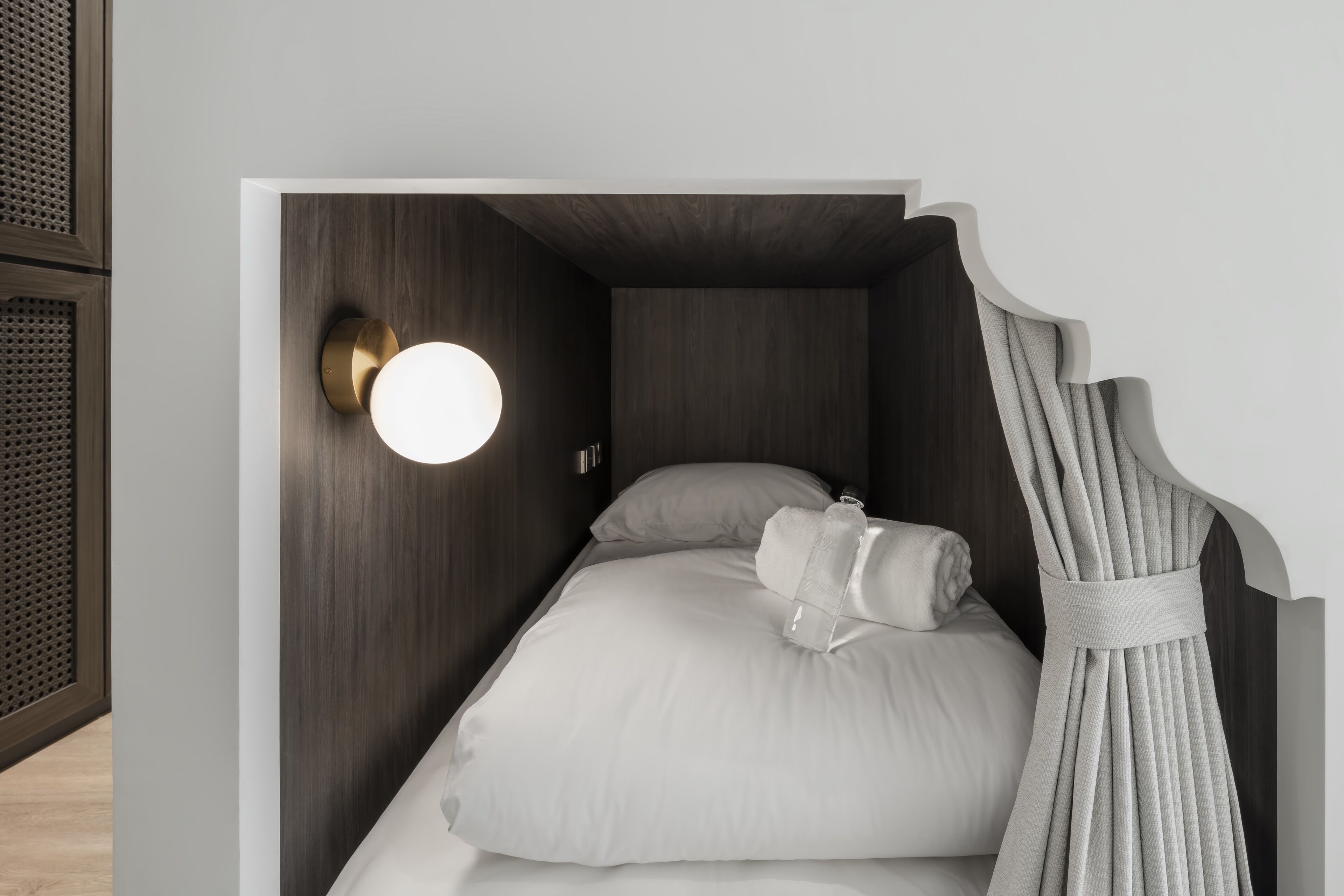 A compact, modern bedroom with a single bed, a round wall-mounted light, and a curtain.