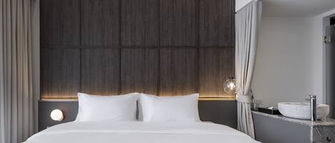 A modern hotel room with a large bed, a wooden headboard, a round wall light, and a bathroom with a vessel sink.