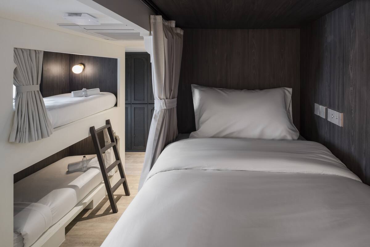 Una habitación de hotel moderna con literas, una cama individual, un tocador con espejo y un pequeño rincón de descanso con una escalera.