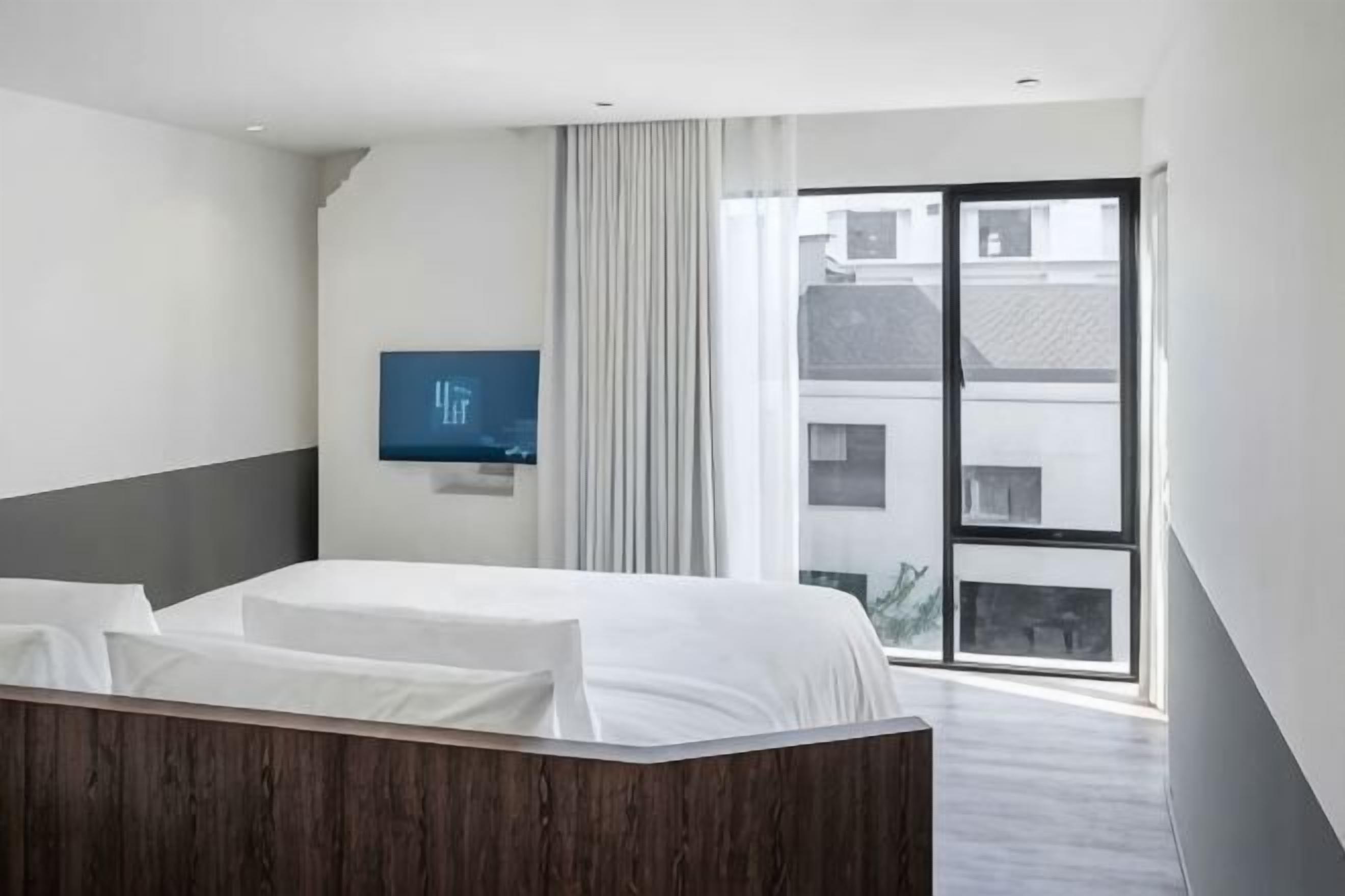 Une chambre d’hôtel moderne équipée d’un lit, d’un téléviseur à écran plat, d’une grande fenêtre avec des rideaux et offrant une vue sur les bâtiments environnants.