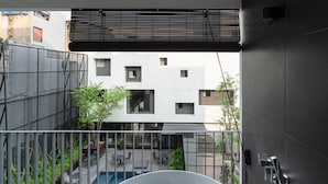 A modern bathroom with a bathtub filled with rose petals, a view of a courtyard with plants and a building, and a balcony with a railing. - Lilit Bang Lumphu Hotel (Bangkok)