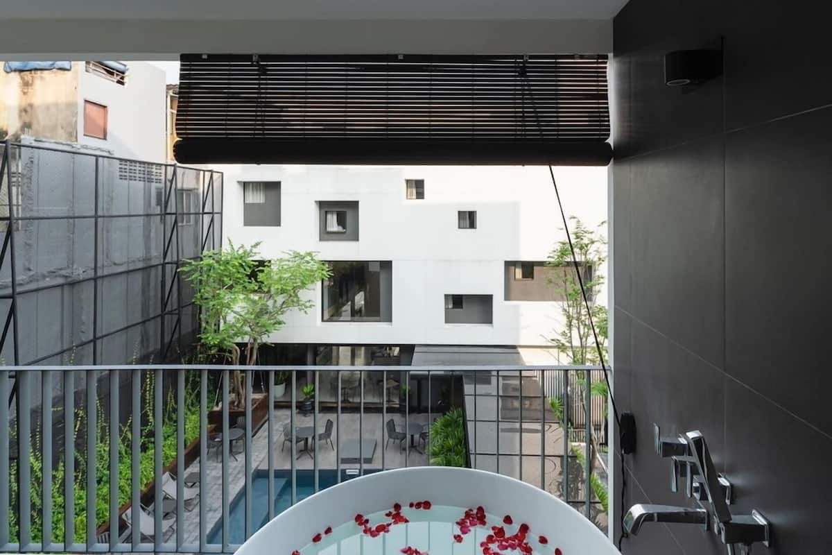 Un baño moderno con una bañera llena de pétalos de rosa, vistas a un patio con plantas y un edificio, y un balcón con barandilla.
