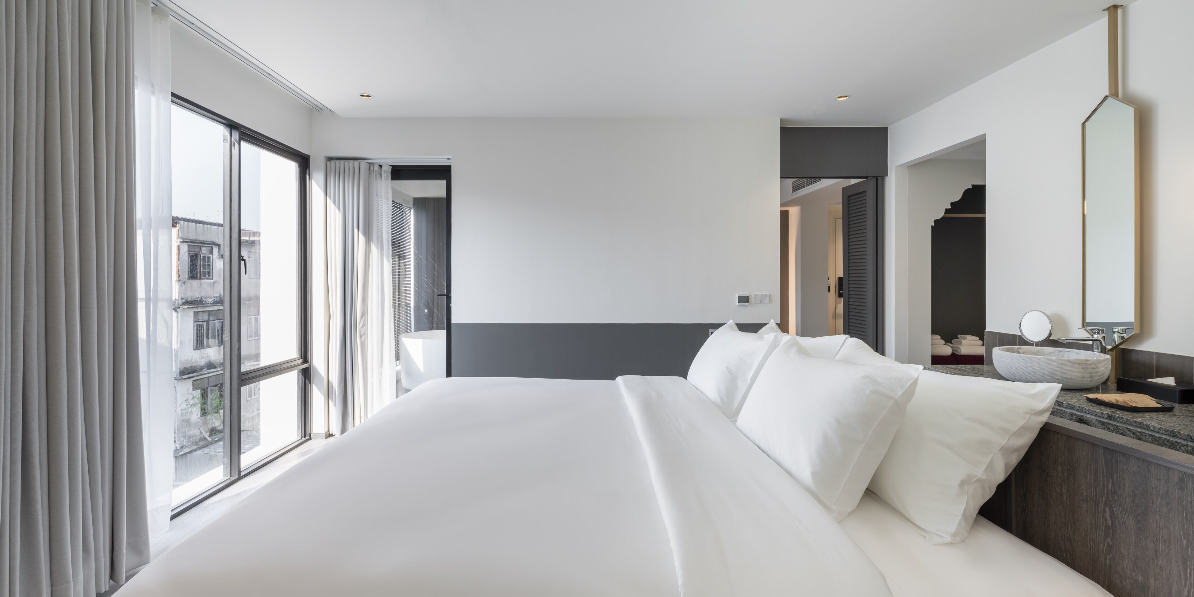 Una habitación de hotel moderna con una cama grande, un baño con lavamanos de mármol y vistas a la ciudad a través de amplias ventanas.