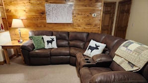 Un salon chaleureux avec un canapé d’angle en cuir marron, deux coussins décoratifs ornés de motifs d’élans, un mur en bois sur lequel est accroché un tableau encadré, et une petite table d’appoint en bois avec une lampe.