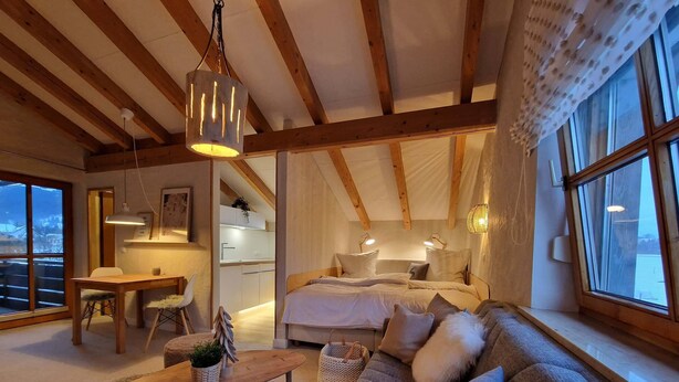 A cozy, well-lit room with a wooden ceiling, a large window, a sofa, a dining table, and a bed with a headboard.