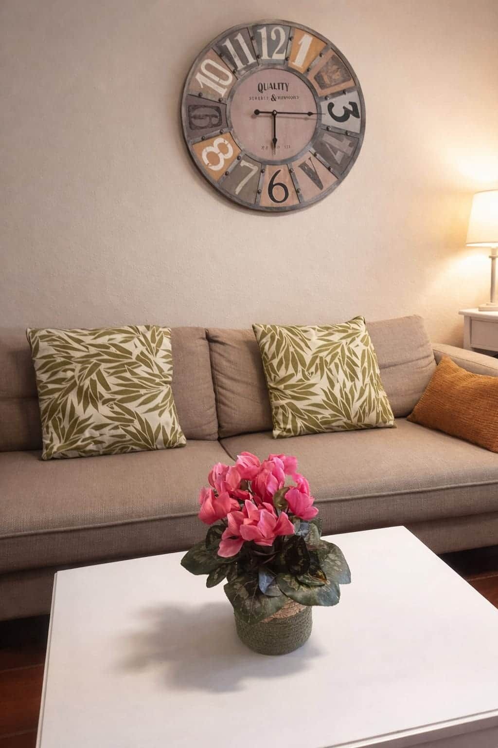 En stue med en beige sofa, mønstrede puder, et hvidt sofabord med en potteplante og et vægur.