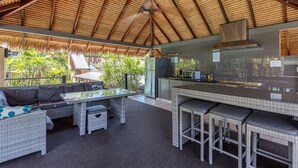 Een buitenkeuken met rieten dak, rieten meubels en uitzicht op tropische begroeiing.