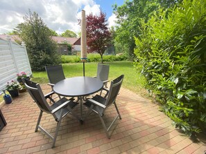 Une terrasse avec une table ronde et quatre chaises, entourée d’un sol en briques et de verdure.