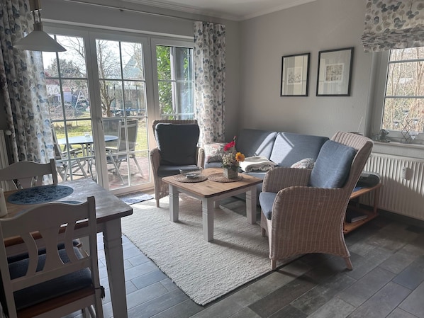 A living room with a sofa, armchair, coffee table, and a dining table with chairs. There is a window with curtains, a radiator, and framed pictures on the wall.