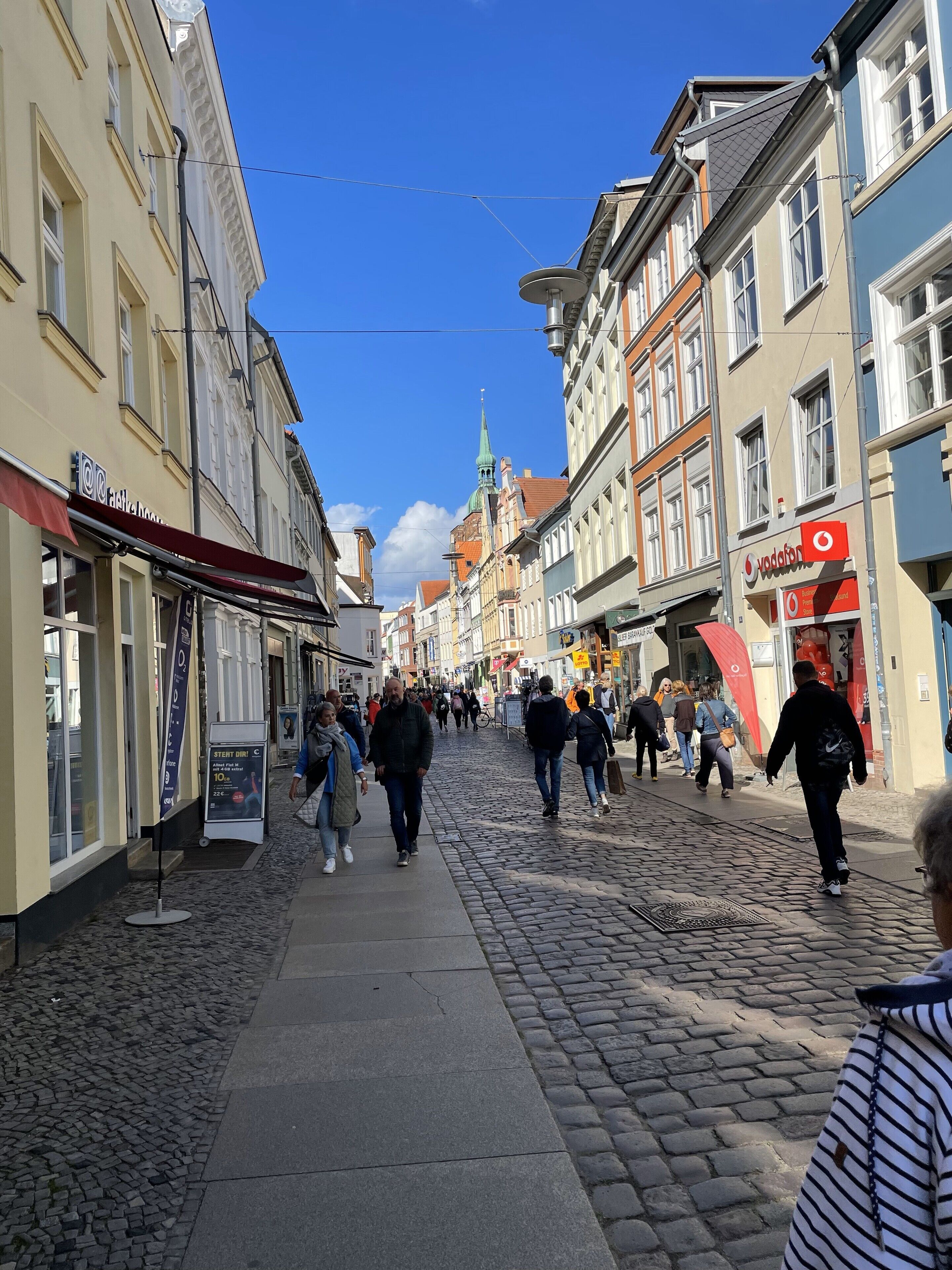 Eine Kopfsteinpflasterstraße gesäumt von historischen Gebäuden, im Hintergrund ein Kirchturm und Fußgänger unterwegs.