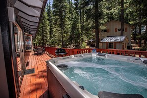 A hot tub surrounded by a wooden deck, with a view of tall pine trees and a building in the background. - Chapel Haus by Hauserman Rental Group (Tahoe City)