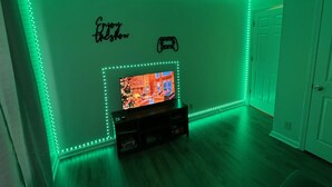 Una habitación con iluminación LED verde a lo largo de las paredes, un televisor sobre un soporte de madera y una puerta a la derecha.