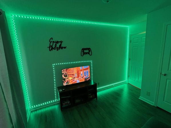 A room with green LED strip lighting along the walls, a television on a wooden stand, and a door to the right.