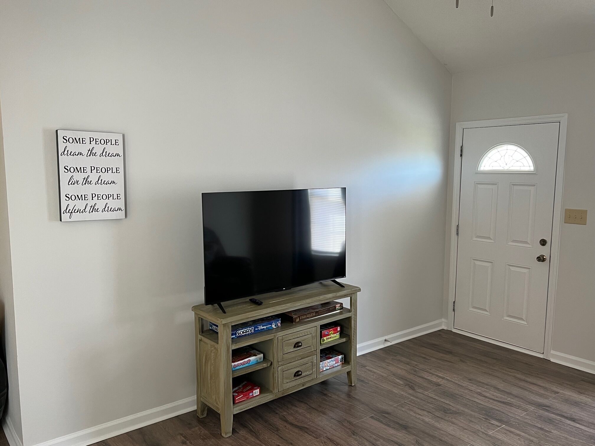A room with a flat-screen TV on a wooden stand, a door with a glass panel, and a wall-mounted sign with motivational text.