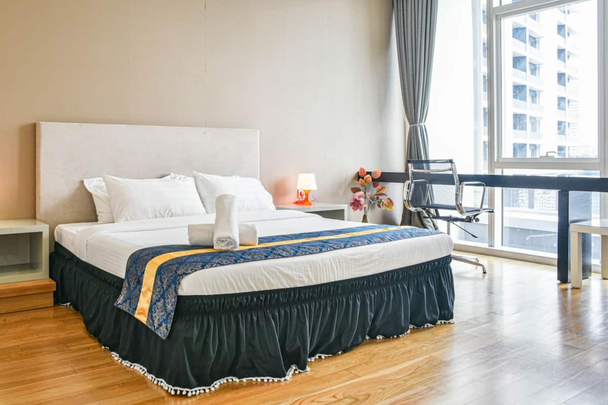 A hotel room with a large bed, a nightstand, a lamp, a chair, and a view of a cityscape through a large window.