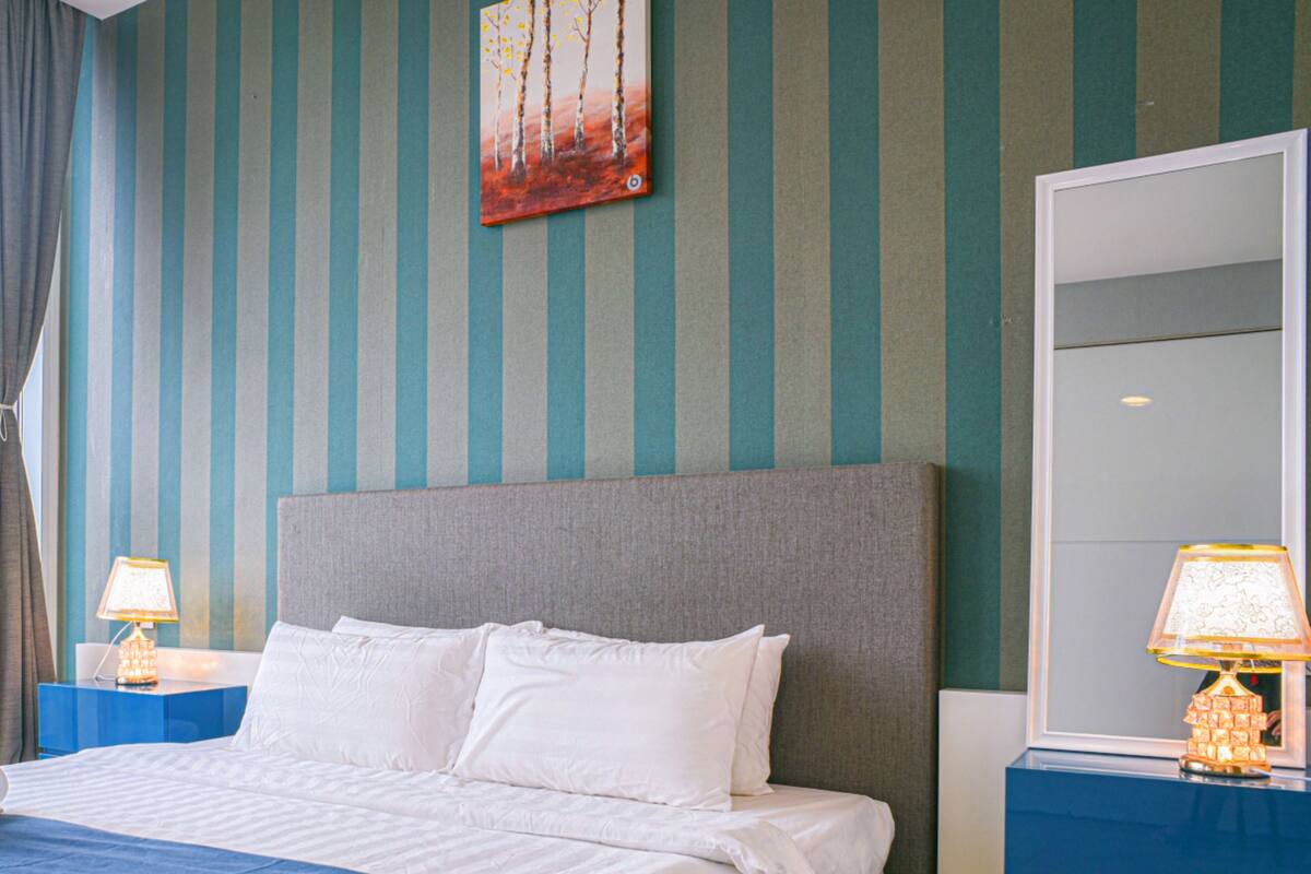A neatly made bed with white linens and a blue accent, a large mirror, and striped walls.