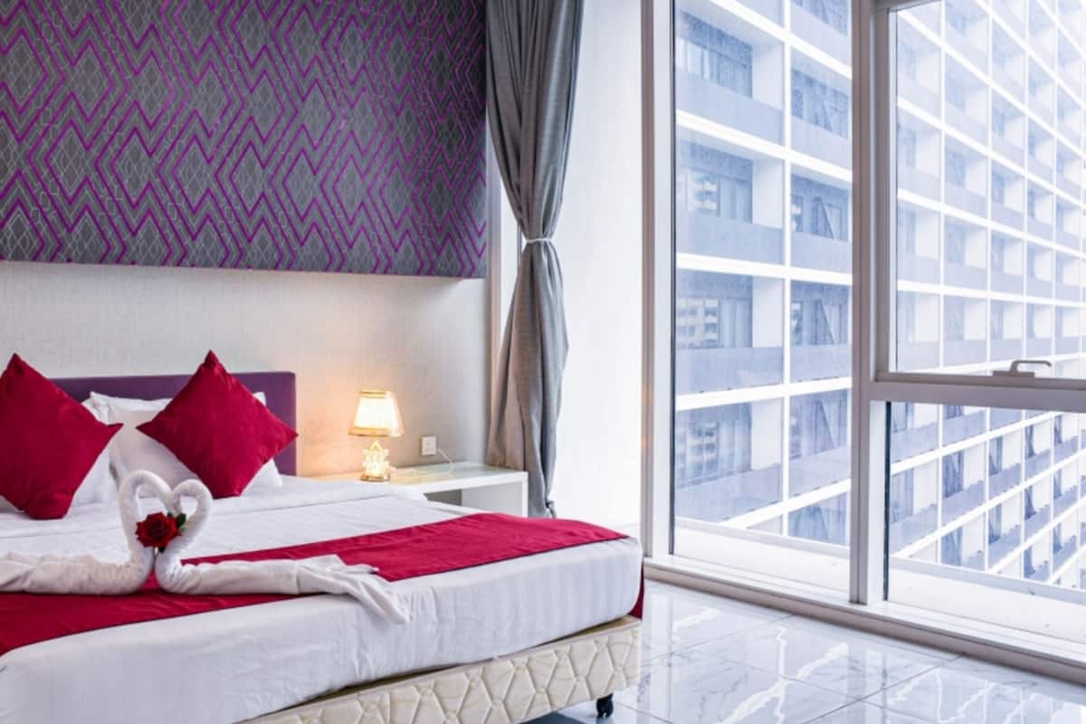 A modern hotel room with a large bed, a nightstand, a lamp, and a view of a cityscape through a large window.