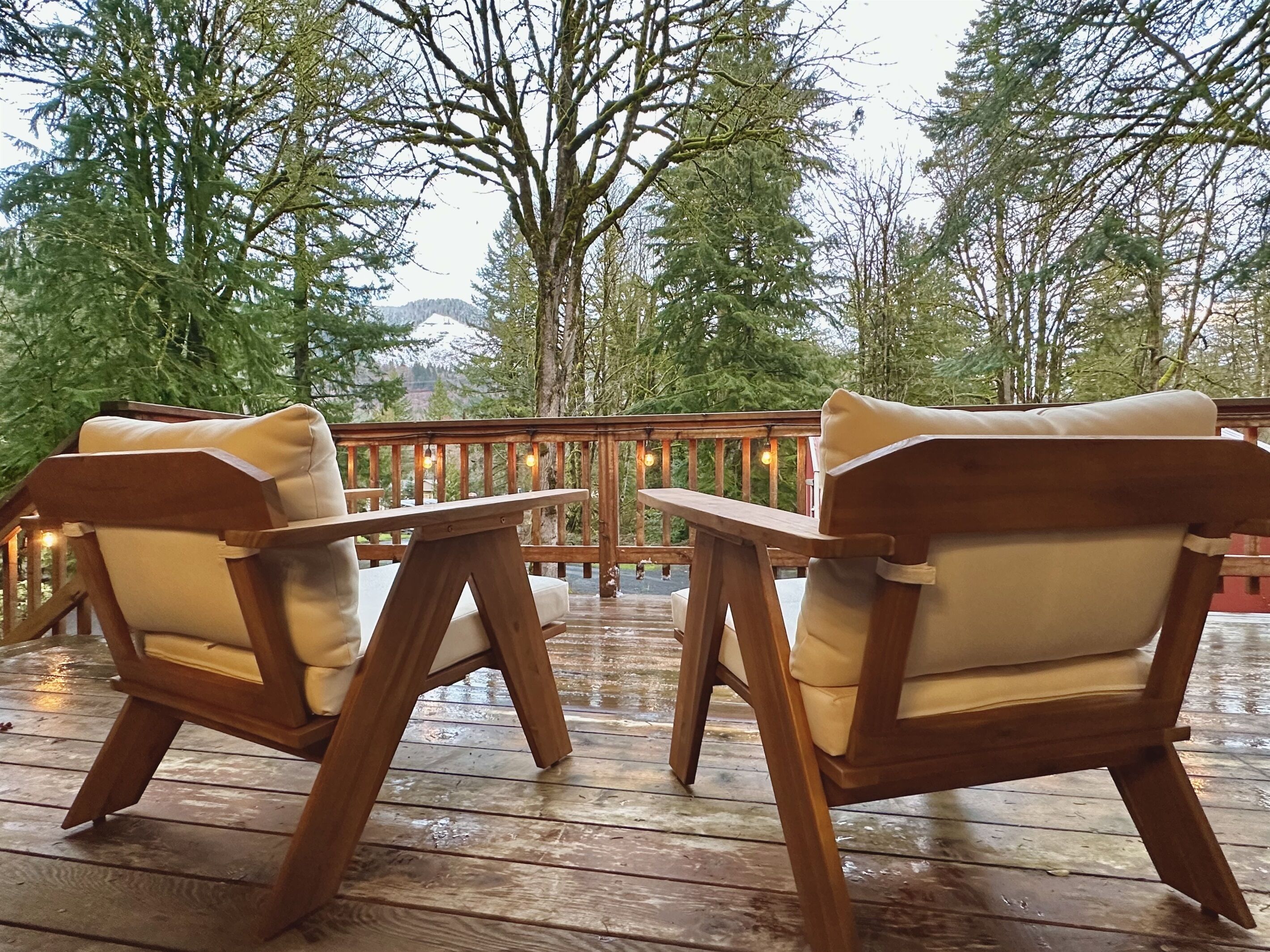 Two wooden chairs with cushions on a wooden deck with a railing, surrounded by trees and a mountain in the background.