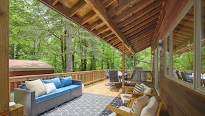 Une terrasse extérieure couverte avec un plafond en bois, un canapé en osier avec des coussins, une table à manger avec des chaises, et une vue sur une zone boisée.