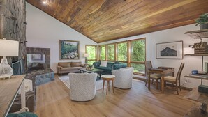 Un vaste espace de vie agrémenté d’une cheminée en pierre, d’un plafond en bois et de grandes fenêtres offrant une vue sur la verdure.