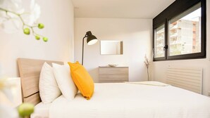 Une chambre moderne comprenant un lit, une table de chevet, une lampe, une commode, un miroir et une fenêtre donnant sur des immeubles.