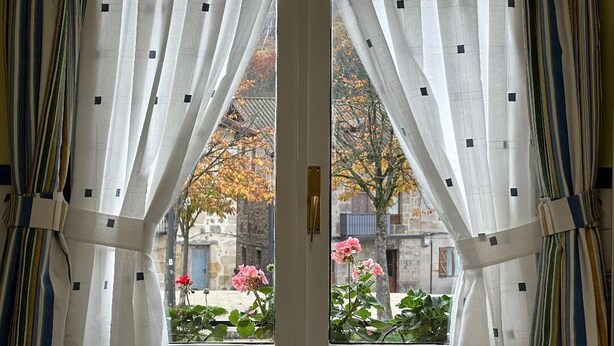 Eine Tür mit zwei Fenstern, jedes mit weißen Vorhängen mit schwarzen Mustern. Draußen Herbstlaub und rosafarbene Blüten an Bäumen.