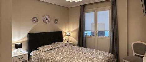 A bedroom with a large bed, a nightstand with a lamp, a chair, and a window with curtains.