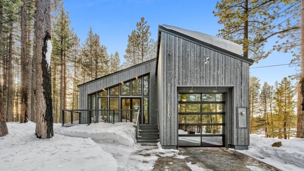 A modern cabin with a large glass door, surrounded by snow and tall trees.