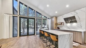 A modern kitchen with a marble countertop, wooden flooring, and large windows offering a view of the outdoors.