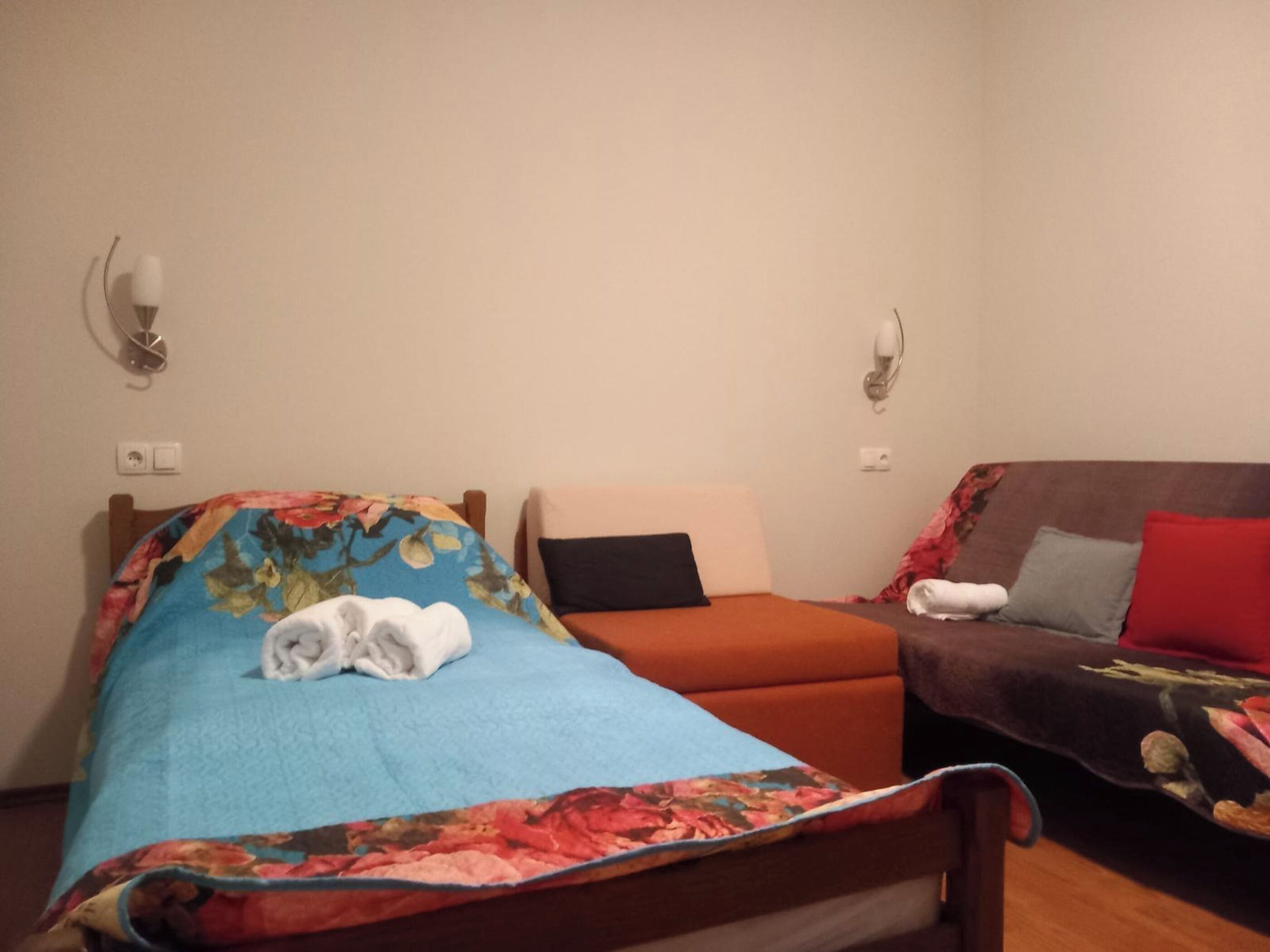 A room with two beds, one with a floral bedspread and the other with a solid color. There are wall-mounted lamps on either side of the beds.