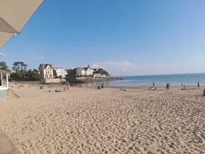 En strand med en stor bygning i baggrunden, sandstrand og klar blå himmel.
