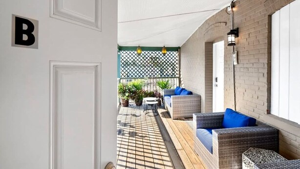 Un espace extérieur abrité, aménagé avec des meubles en osier et des coussins bleus, un plancher en bois et un mur en briques.