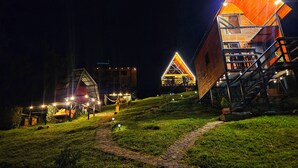 A series of uniquely designed cabins with sloped roofs and wooden exteriors, arranged on a grassy hillside with a brick pathway leading through them. - Hakuna Matata glamping (Monguí)