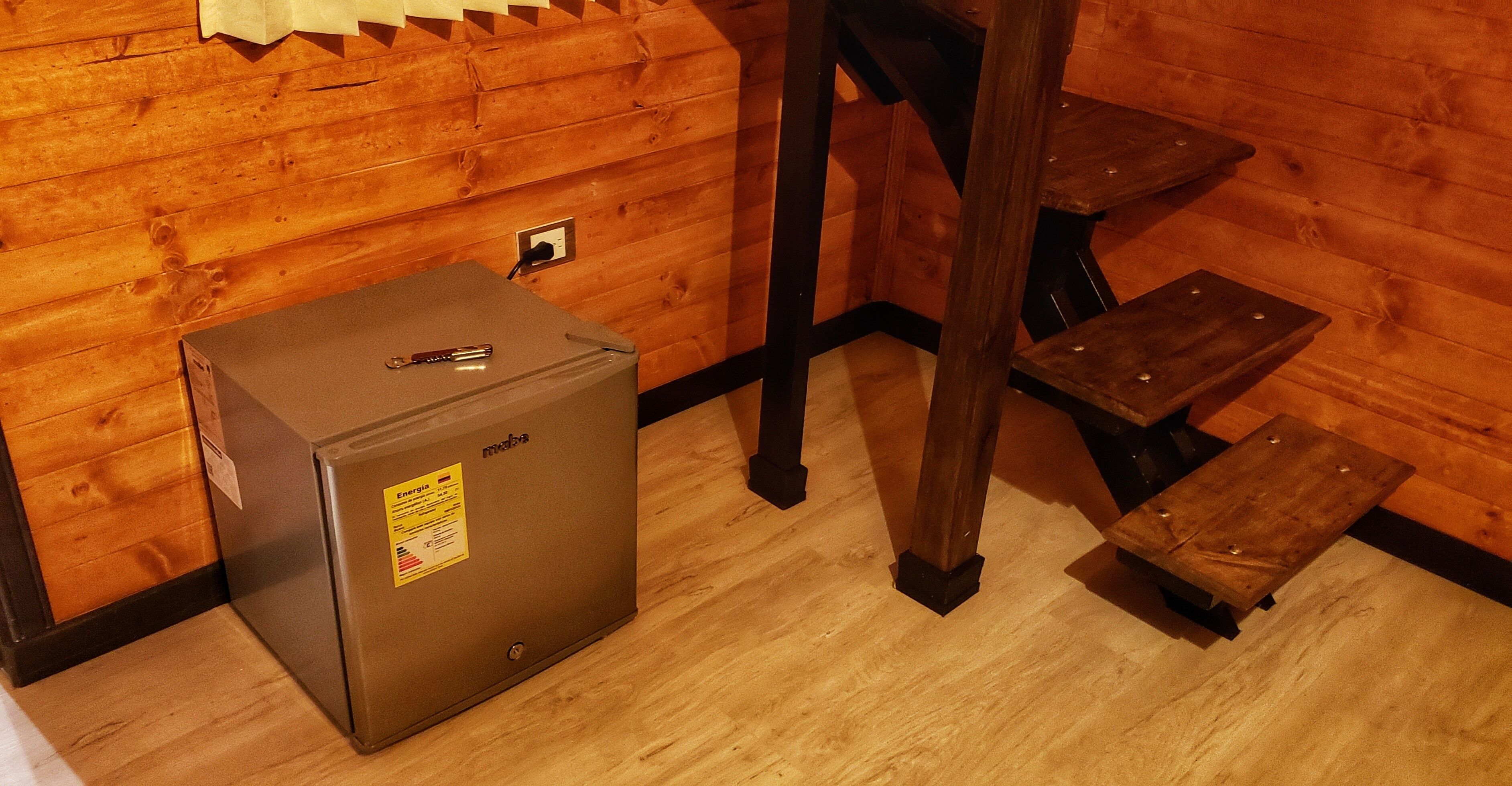 Un petit local technique aux finitions métalliques, comprenant un petit réfrigérateur, une prise de courant et un tabouret en bois.