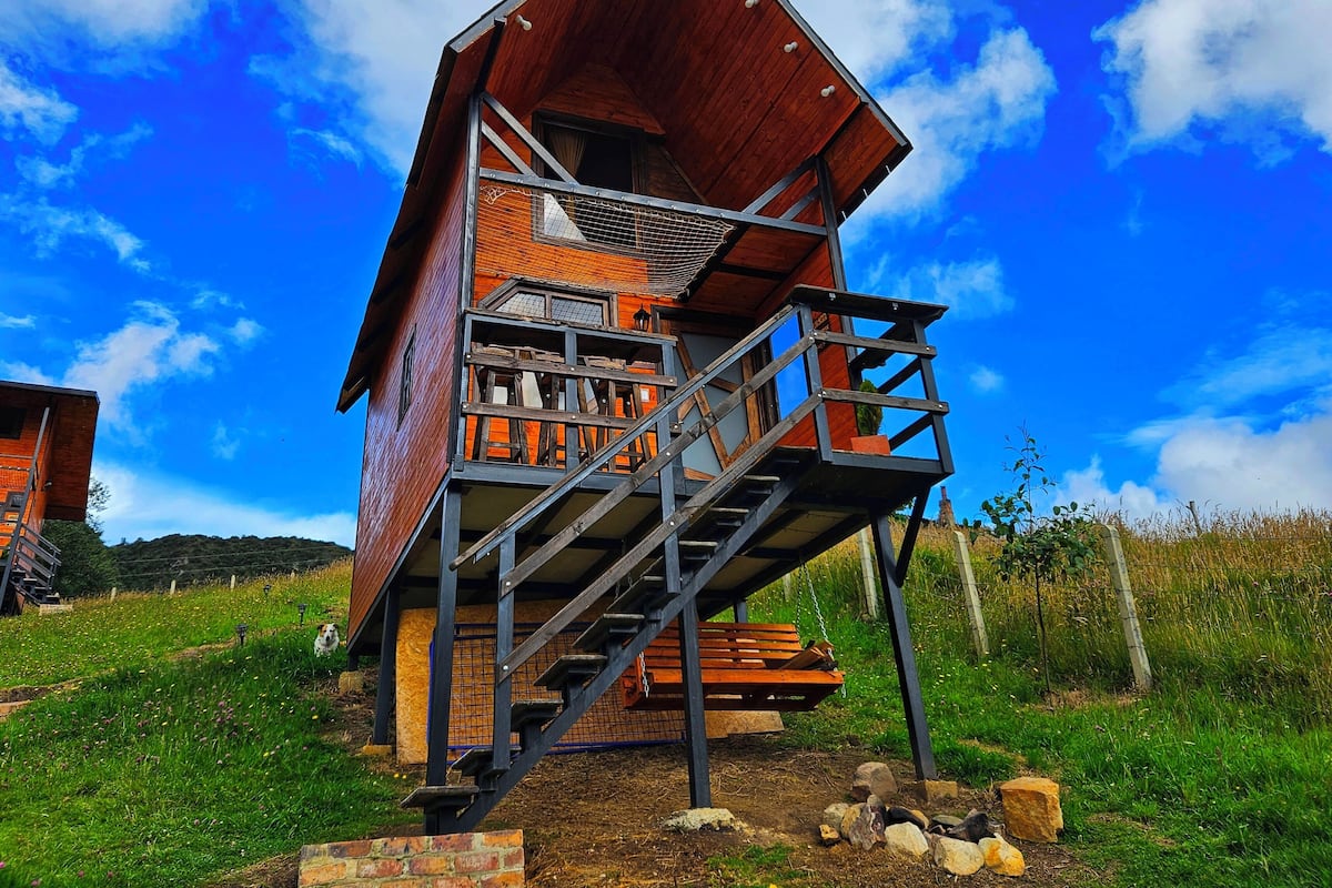 Una cabaña de madera con balcón y escaleras, rodeada por un campo cubierto de césped y un cielo despejado.