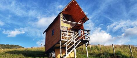 A wooden cabin with a balcony and stairs, set in a grassy field with a clear sky.