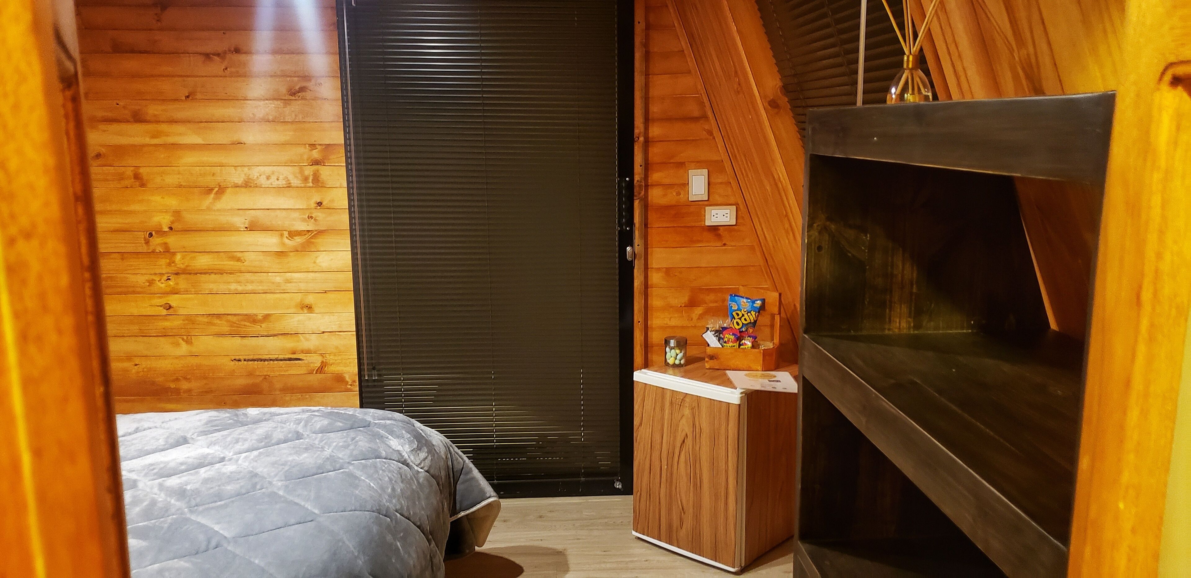 A compact room with a bed, a small wooden cabinet with snacks, a TV, and a fireplace.