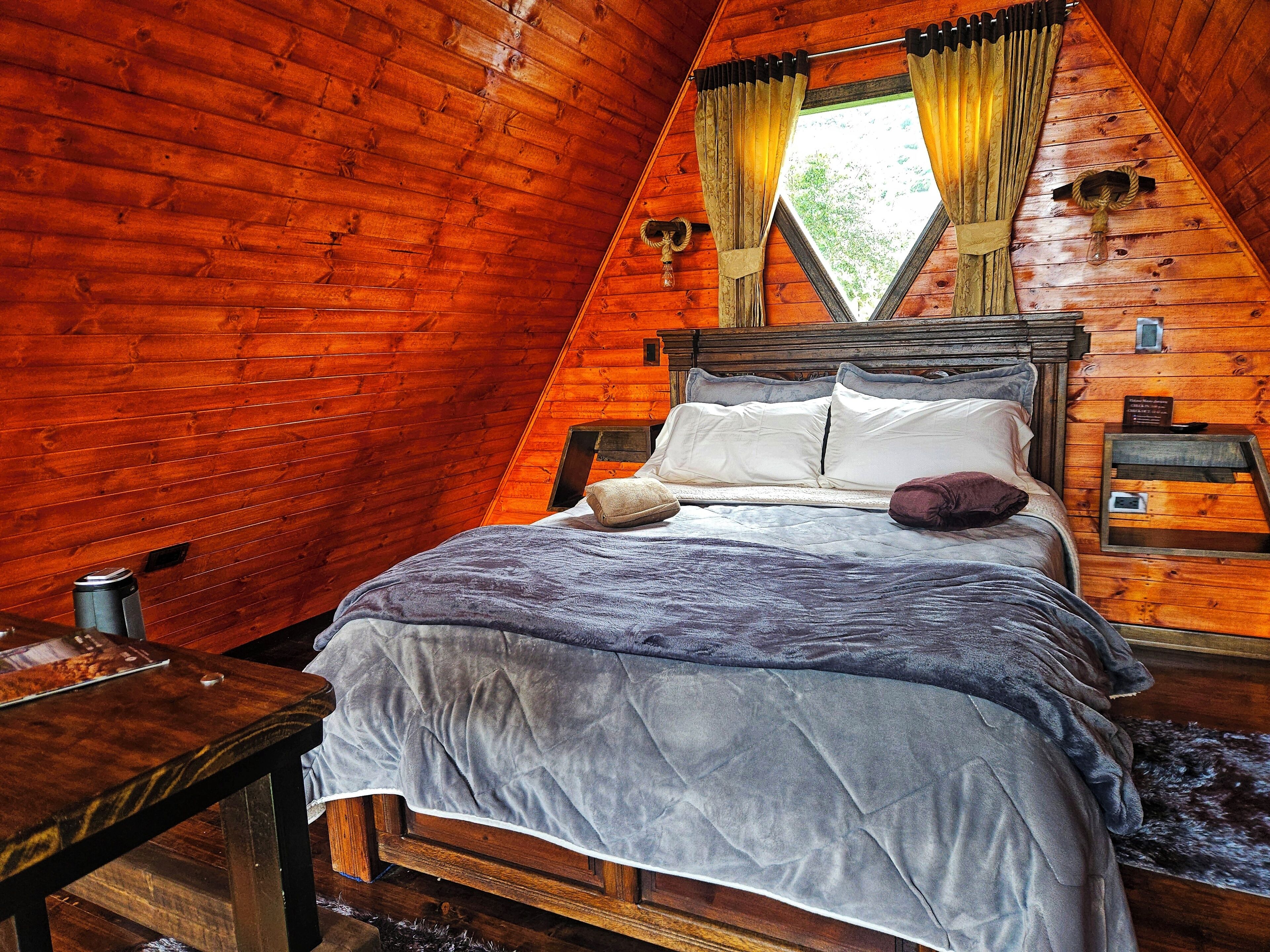 Un dormitorio acogedor tipo cabaña de madera con cama, mesita de noche, ventana con cortinas y pared de madera.