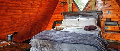 Un dormitorio acogedor tipo cabaña de madera con cama, mesita de noche, ventana con cortinas y pared de madera.