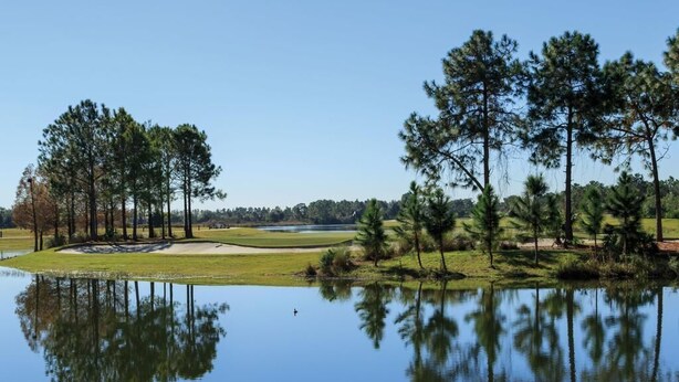 Rauhallinen golfkenttä, jonka yhteydessä on tyyni järvi, jonka pinnasta heijastuvat puut ja kirkas taivas.