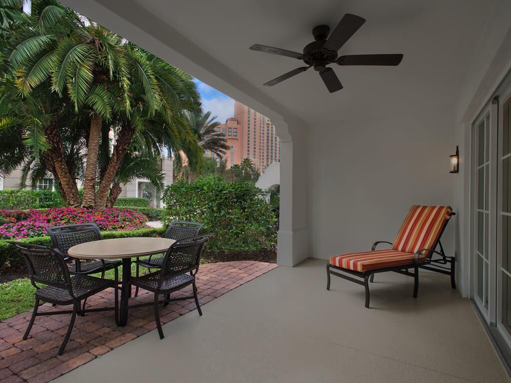 A covered patio with a table and chairs, a striped lounge chair, a ceiling fan, and a view of palm trees and buildings.