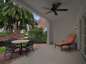 Portico coperto con tavolo e sedie, una poltrona da relax a righe, una ventola a soffitto e vista su palme e edifici.