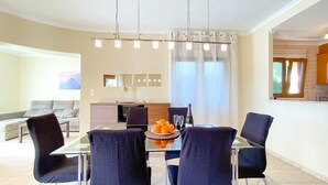 A modern dining area with a glass table, dark chairs, and a fruit bowl. There is a kitchen in the background with a window and a view of greenery. - Quiet and cozy villa near the beach (Denia)