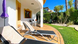 A covered outdoor seating area with lounge chairs and a table, surrounded by a well-maintained lawn and palm trees. - Quiet and cozy villa near the beach (Denia)