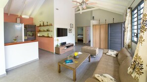 Een ruime woonkamer met een bank, salontafel en televisie. Er is een keuken met een koelkast en een eettafel. De kamer heeft een hoge plafond met een ventilator en grote ramen.